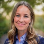 Dr. Heinsberg head shot while wearing a blazer in the outdoors with green leaves behind her.